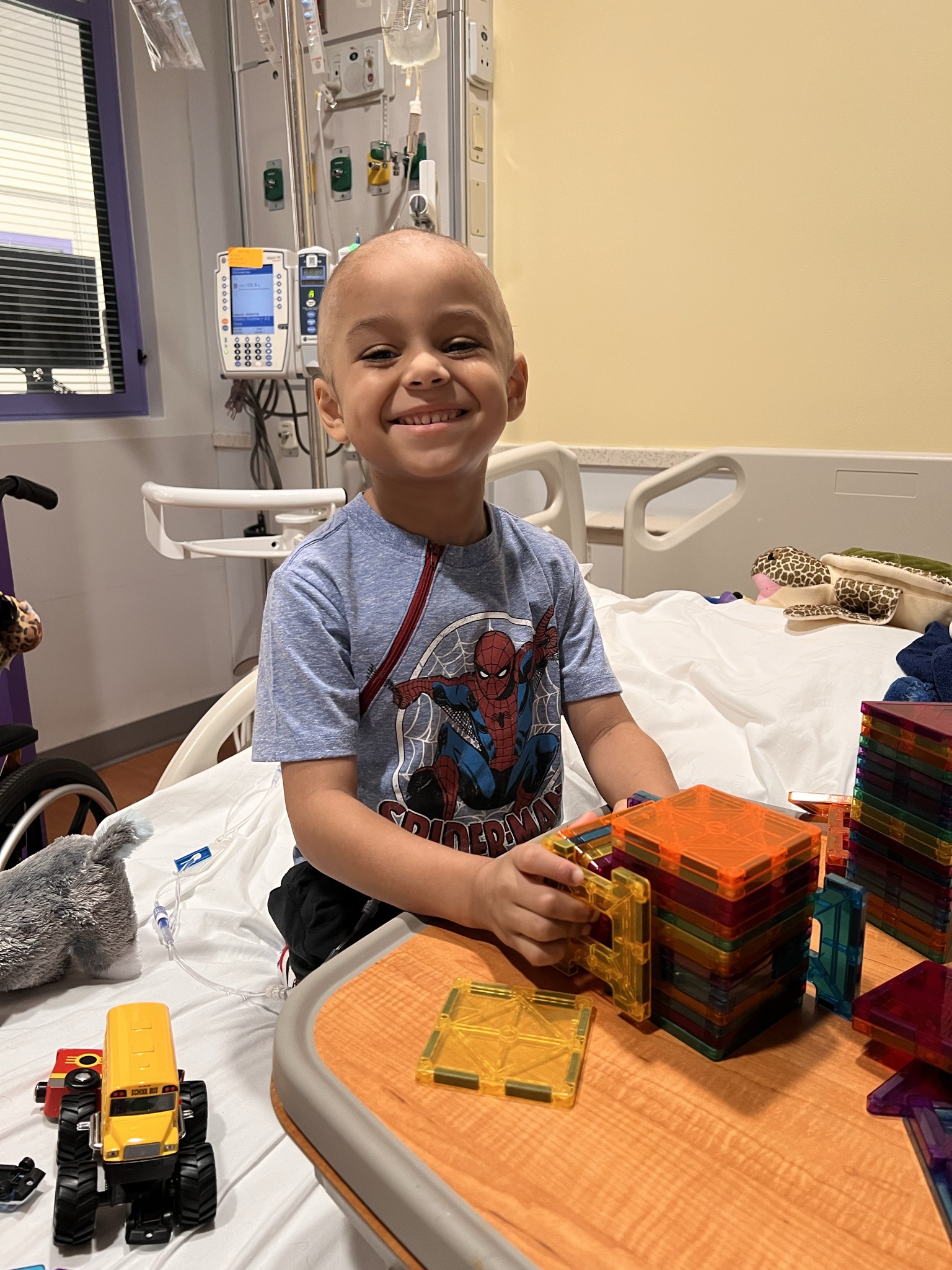 Kelvon wearing a Spider-Man shirt and smiling from his hospital bed while building structures with colorful magnetic tiles on a bedside table.
