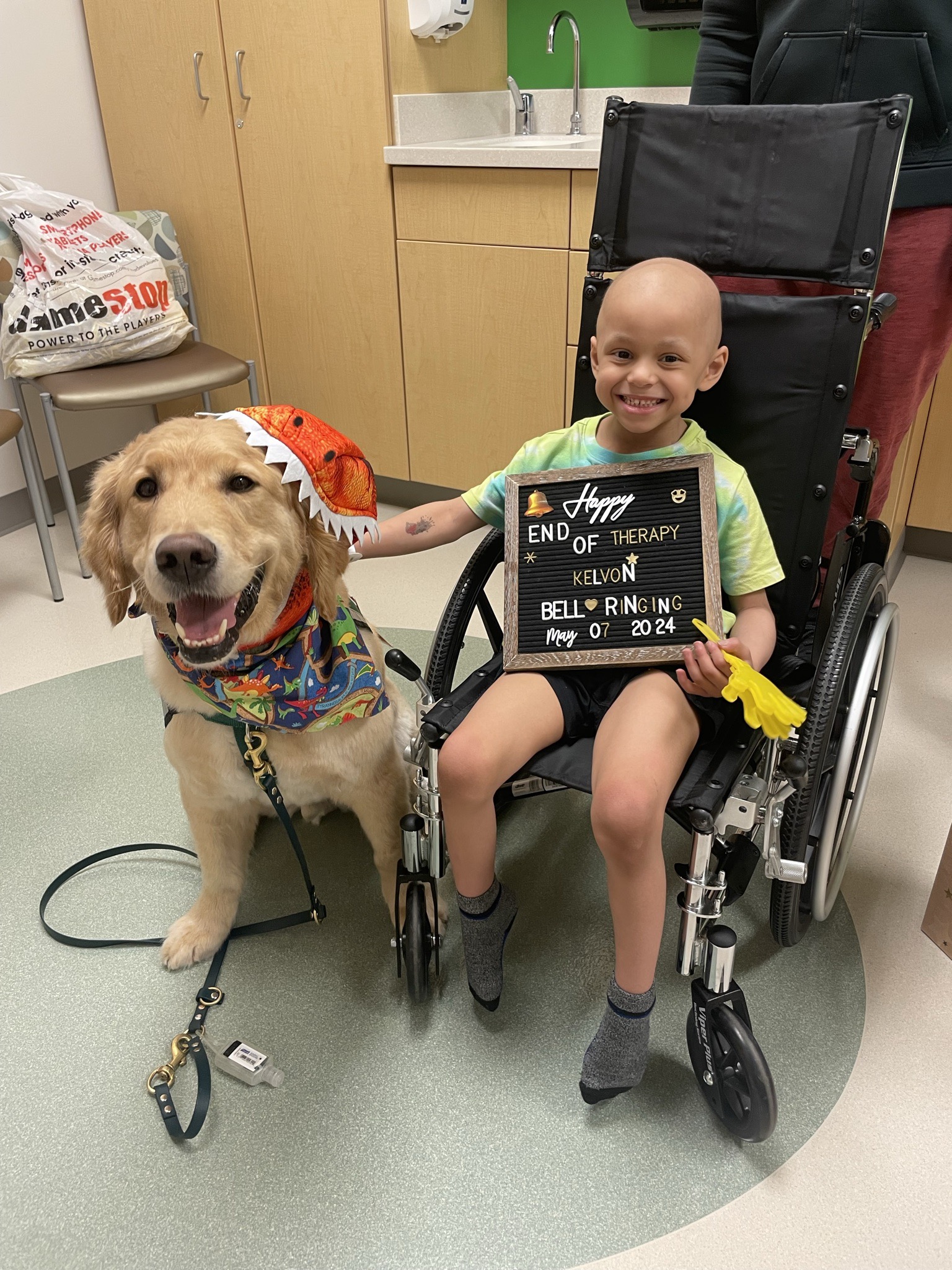 Kelvon smiling in his wheelchair holding a letter board celebrating the end of therapy and bell ringing on May 7, 2024, next to a therapy dog in a dinosaur costume.
